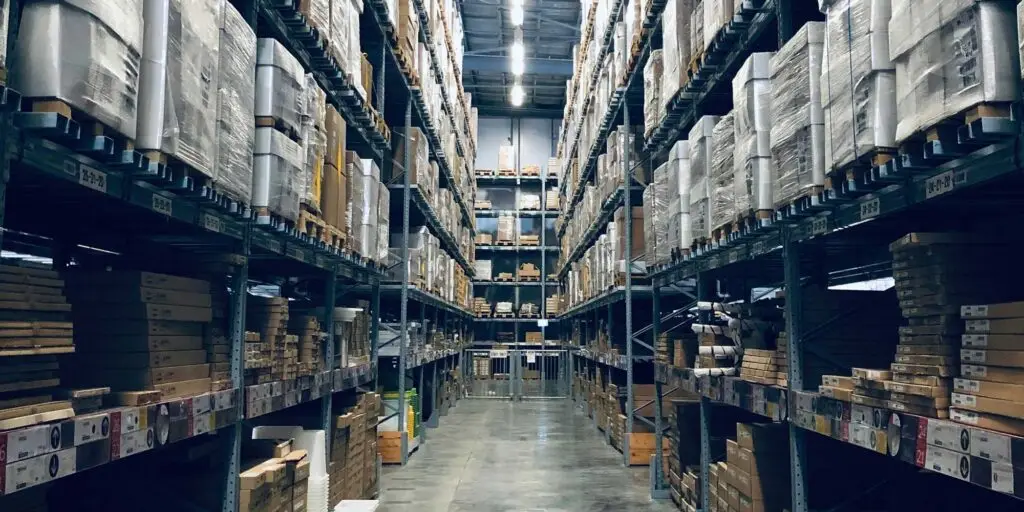 Høye pallereoler fulle av innpakkede varer står langs en bred lagergang, med esker stablet nederst og oppgjør fra nettbutikker i bakgrunnen.