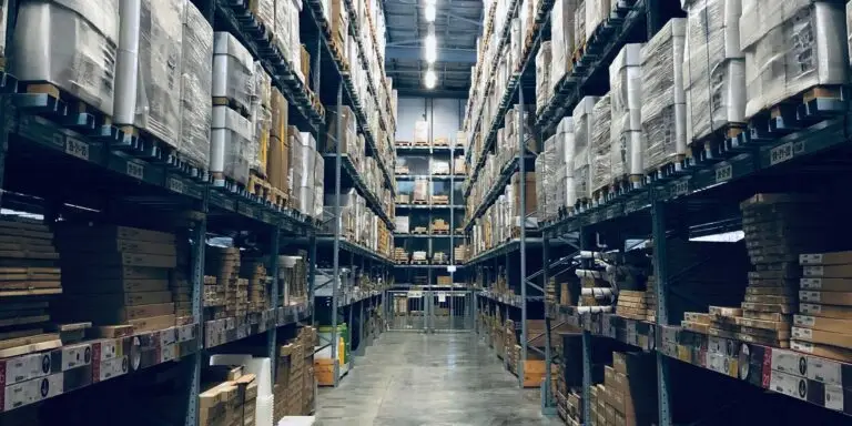 Høye pallereoler fulle av innpakkede varer står langs en bred lagergang, med esker stablet nederst og oppgjør fra nettbutikker i bakgrunnen.
